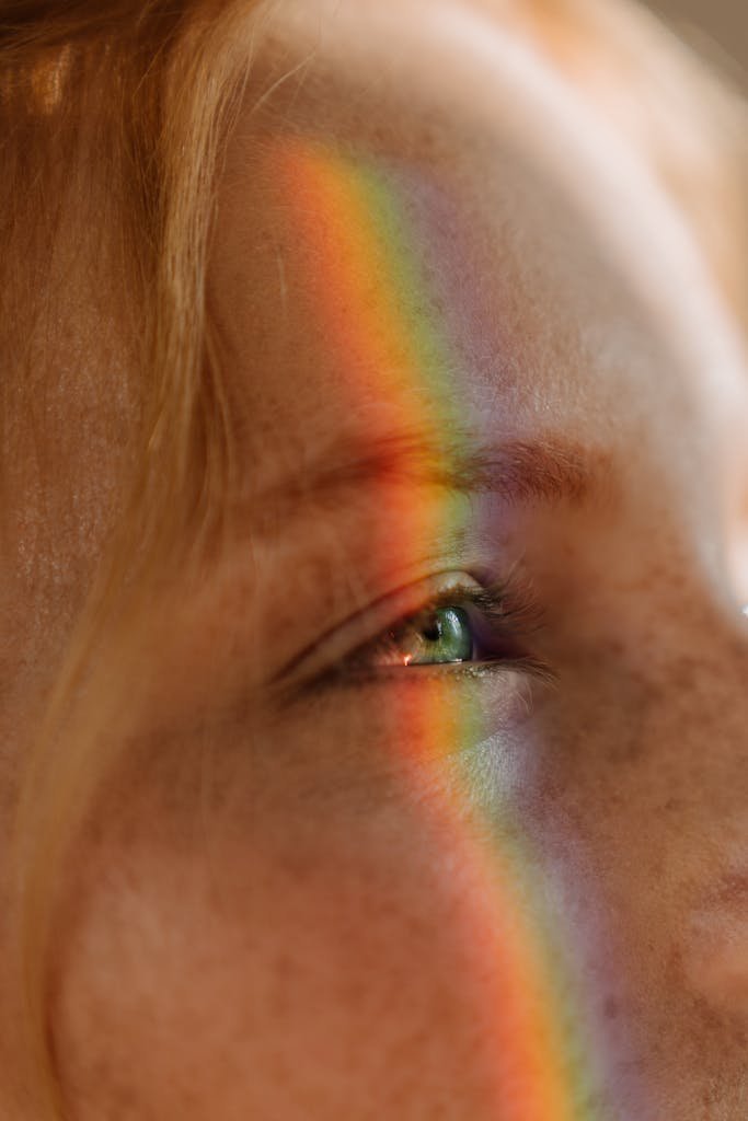 Artistic close-up of a freckled face with vibrant rainbow light across the eye, conveying beauty and diversity.