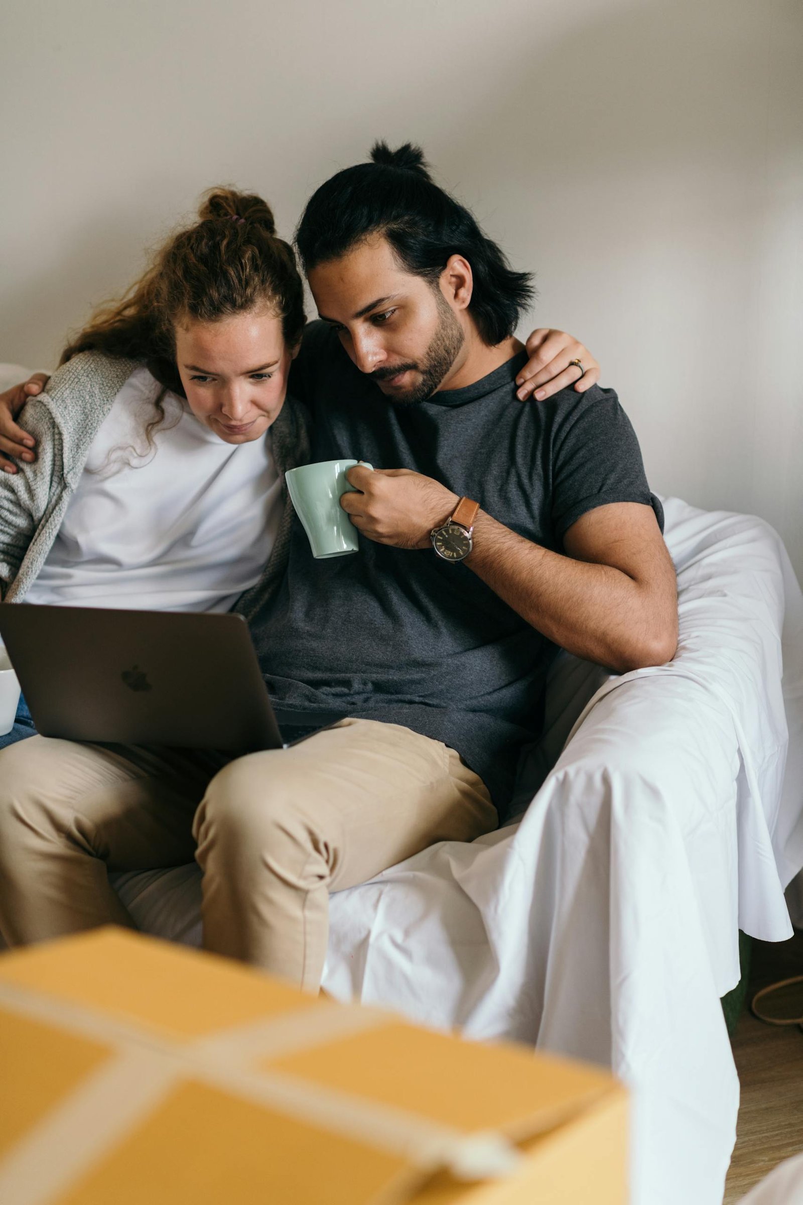 Young couple embracing while online shopping on laptop, sipping coffee, surrounded by moving boxes at home.